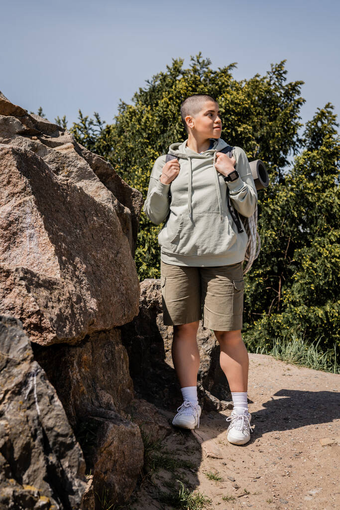Young short haired female tourist with fitness tracker and backpack looking away while standing near stones with nature and blue sky at background, exploring new horizons, Translation of tattoo: love - Photo, Image