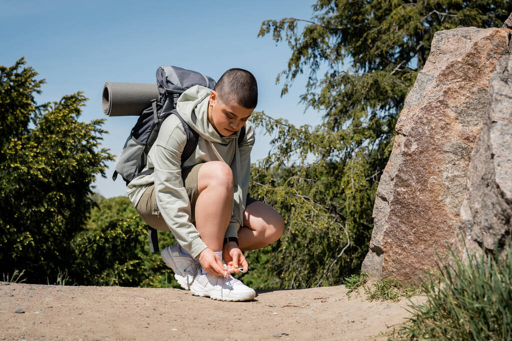 Young short haired female traveler with backpack and fitness mat tying shoelace on sneaker and standing near stones on path with nature at background, trekking through rugged terrain - Photo, Image