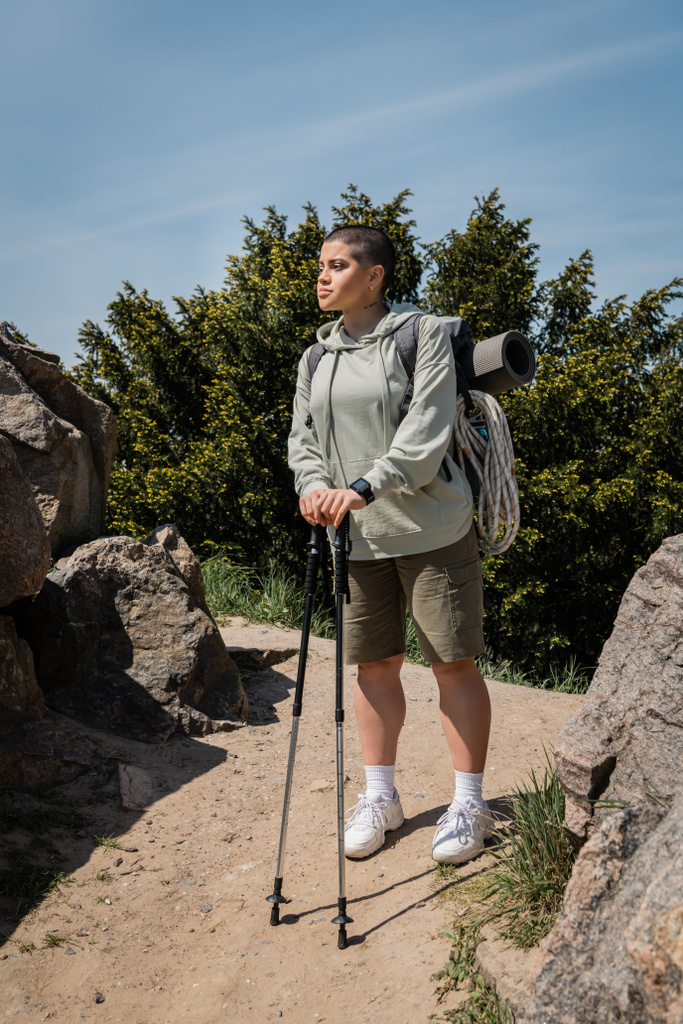 Młody, krótkowłosy i wytatuowany wędrowiec żeński z fitness-trackerem i plecakiem ze sprzętem podróżniczym trzymającym kijki trekingowe stojąc na ścieżce z kamieniami, wędrując po nierównym terenie - Zdjęcie, obraz