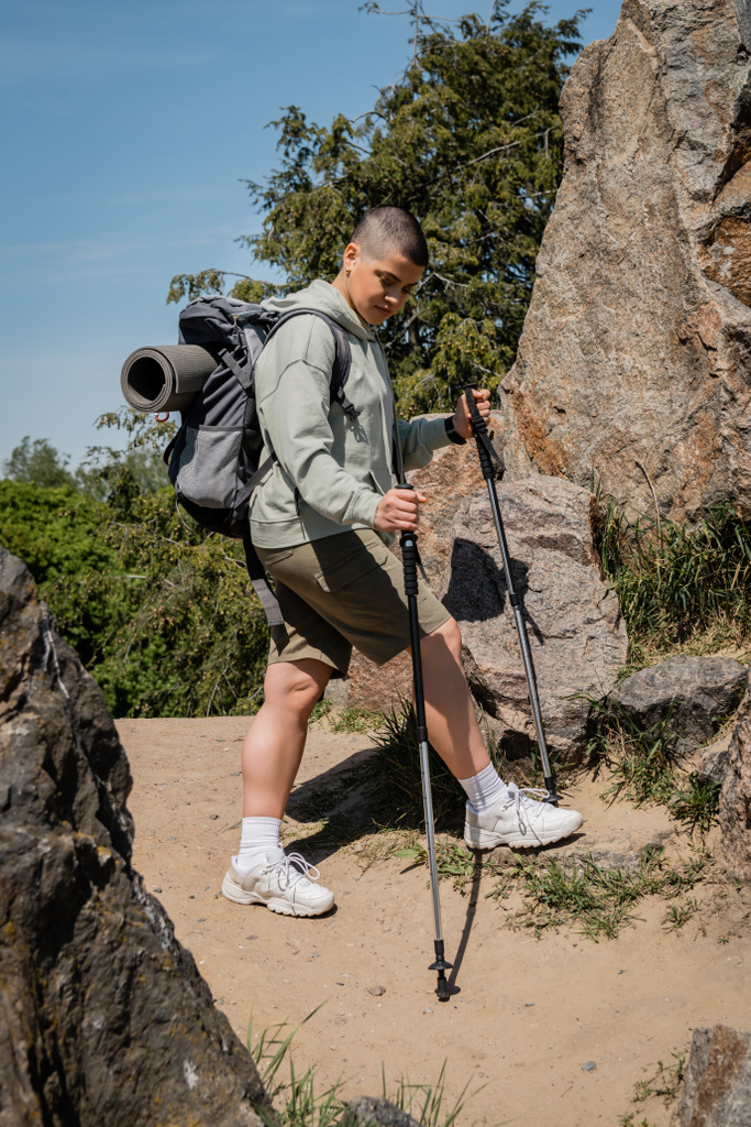 Young short haired female hiker with backpack holding trekking poles and looking at path while standing between stones with blue sky at background, trekking through rugged terrain, summer - Photo, Image