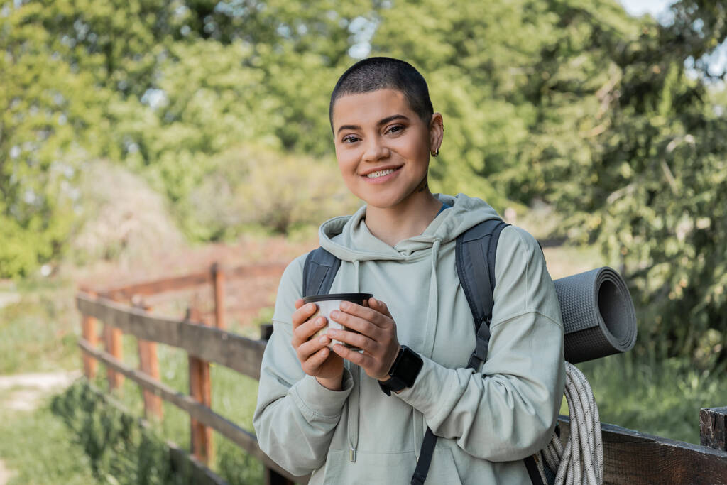 Cheerful young short haired female hiker with fitness tracker and backpack holding thermos cup and looking at camera with landscape at background, finding serenity in nature, summer - Photo, Image