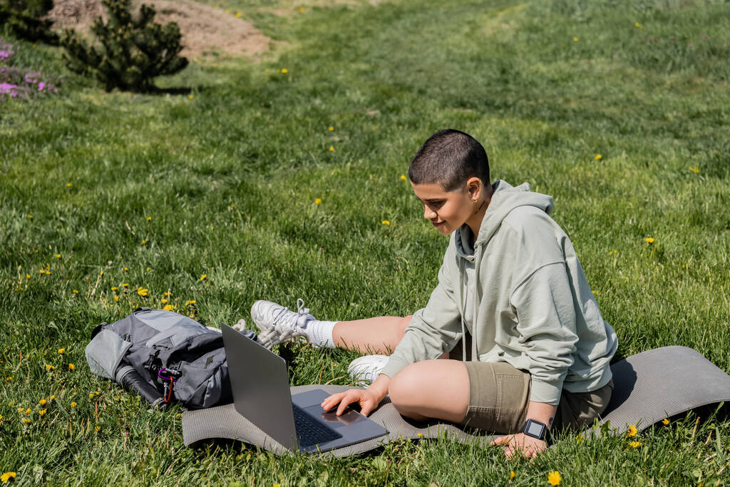 Junge kurzhaarige Touristin in lässiger Kleidung mit Laptop, während sie auf einer Fitnessmatte in der Nähe eines Rucksacks auf einer Rasenfläche mit Blumen sitzt und Gelassenheit in der Natur findet, Sommer - Foto, Bild