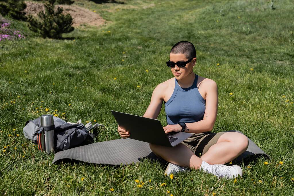Junge kurzhaarige Touristin mit Sonnenbrille und Laptop auf Fitnessmatte neben Rucksack und Thermoskanne auf Rasen mit Blumen, Ruhe in der Natur finden, Sommer, digitaler Nomade  - Foto, Bild