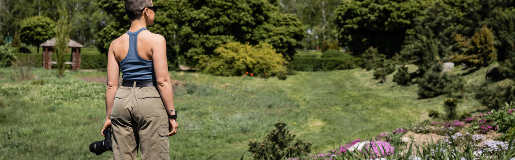 Seitenansicht einer jungen kurzhaarigen Touristin mit Sonnenbrille, die eine Digitalkamera hält und in der Nähe von Blumen steht, mit einer malerischen Landschaft im Hintergrund, die sich mit dem Naturkonzept verbindet, Banner  - Foto, Bild