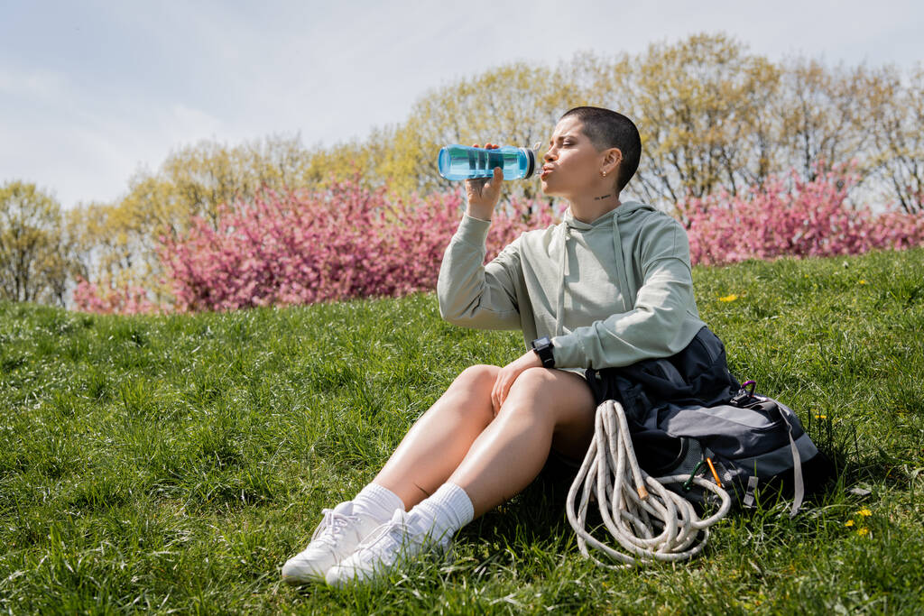 Nuori lyhytkarvainen ja tatuoitu naispuolinen reppumatkailija, jolla on älykello juomaveden kanssa istuessaan lähellä reppua nurmikolla, jossa luonto on taustalla, ja joka kulkee maiseman läpi - Valokuva, kuva