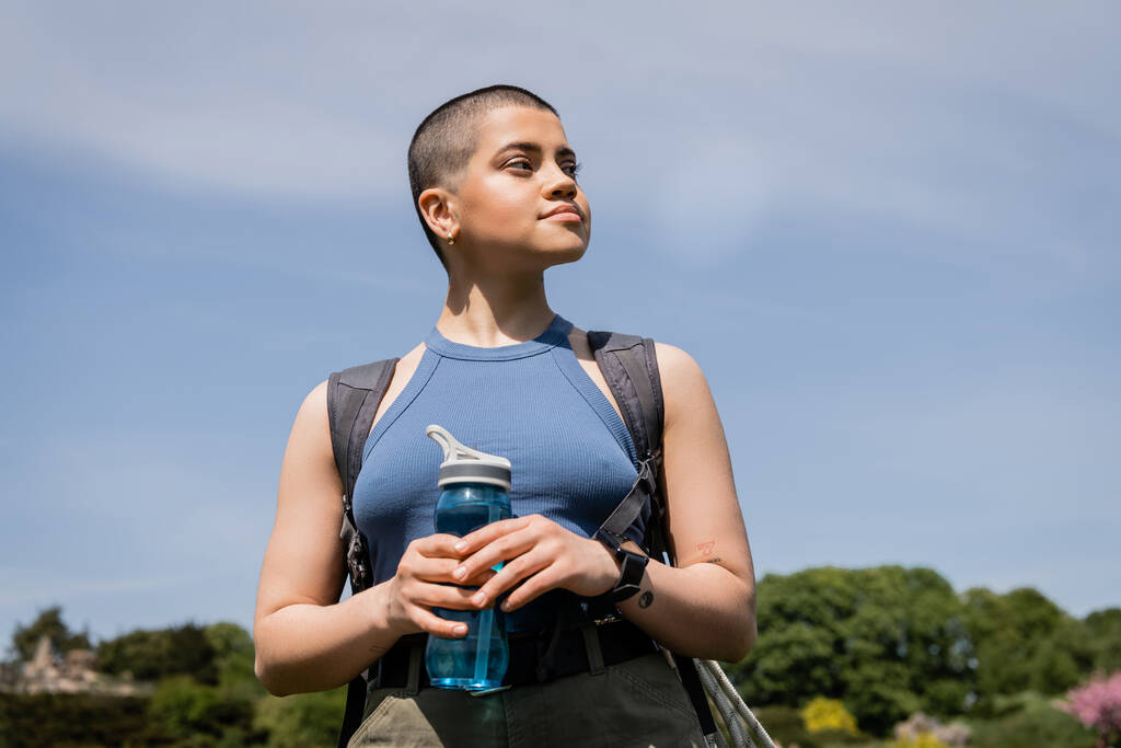 Junge tätowierte und kurzhaarige Wanderin mit Rucksack und Smartwatch, die wegschaut und im Hintergrund eine Sportflasche mit blauem Himmel hält, bahnbrechend durch malerische Landschaft, Sommer - Foto, Bild