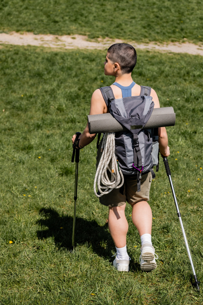Seitenansicht einer jungen kurzhaarigen Touristin mit Rucksack und Reiseausrüstung, die Trekkingstöcke hält, während sie im Hintergrund auf einer Wiese spaziert, Einzelwanderungskonzept, Sommer - Foto, Bild