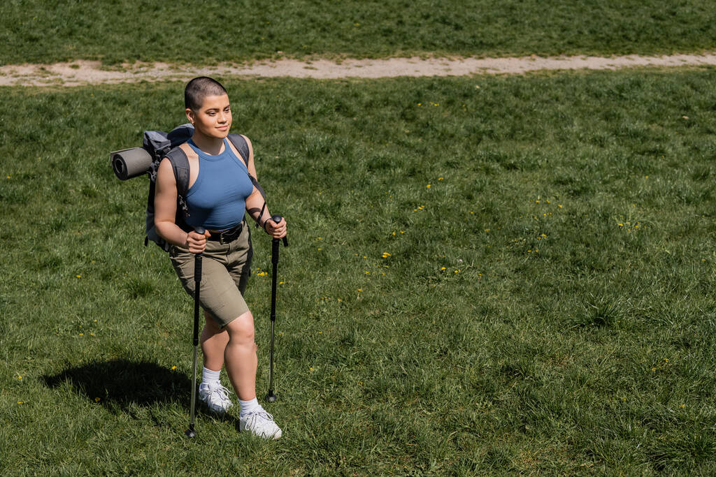 Vista de ángulo alto de sonriente joven excursionista femenina de pelo corto con mochila sosteniendo bastones de trekking y caminando sobre césped cubierto de hierba en el fondo, concepto de viaje de senderismo en solitario, verano - Foto, imagen