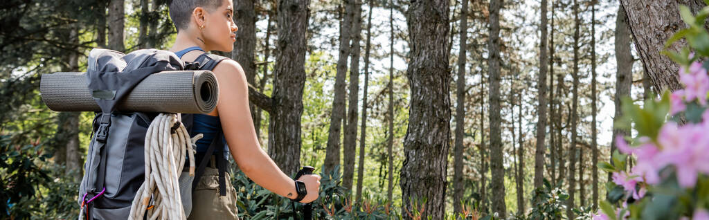 Vista lateral de la joven viajera femenina de pelo corto y tatuada con mochila sosteniendo el poste de trekking y de pie en el verde bosque de verano, reconectando con usted mismo en el concepto de la naturaleza, bandera  - Foto, imagen