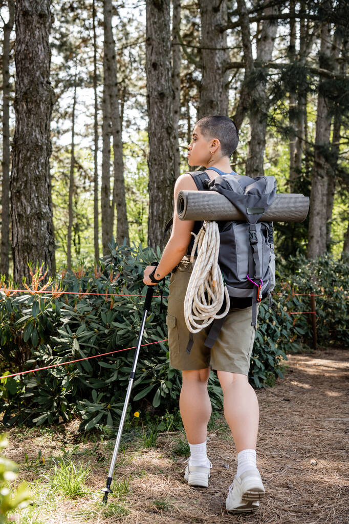 Vista lateral de una joven viajera de pelo corto con mochila sosteniendo poste de trekking y senderismo en el verde bosque de verano, reconectando con usted mismo en concepto de naturaleza - Foto, Imagen