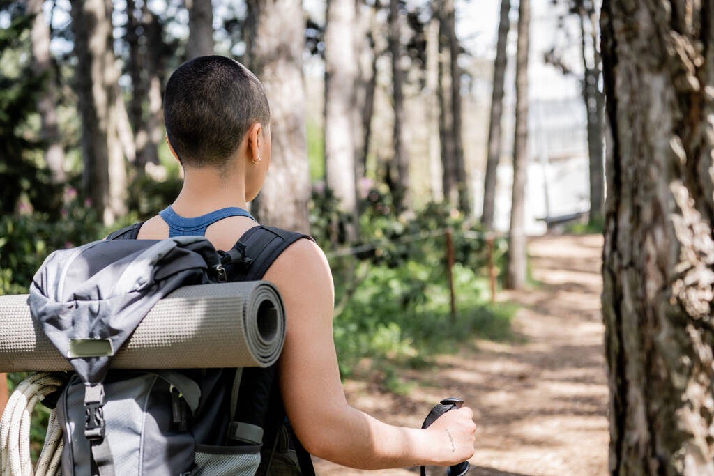 Rückenansicht einer jungen, kurzhaarigen und tätowierten Touristin mit Rucksack und Fitnessmatte, die beim Wandern im verschwommenen grünen Wald Trekking-Stöcke hält, Übersetzung des Tattoos: Liebe - Foto, Bild