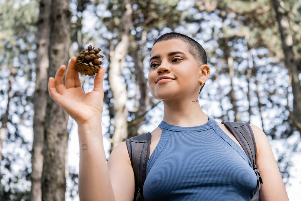 Low angle view of young tattooed and short haired female tourist with backpack holding pine cone while standing in blurred summer forest, hiking for health and wellness concept  - Photo, Image