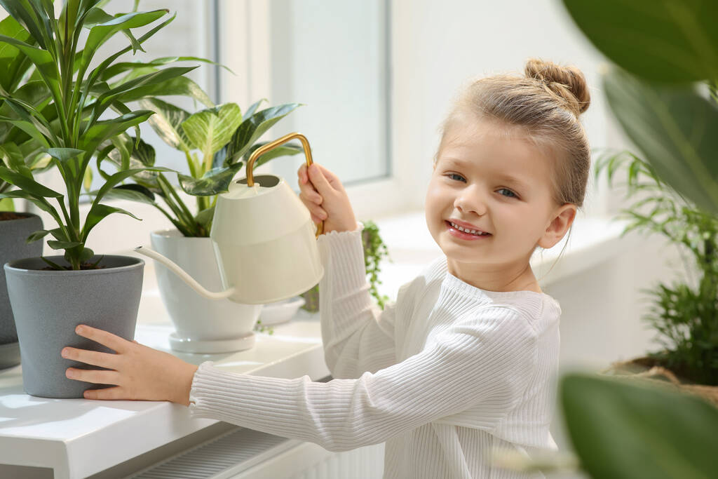 Милая маленькая девочка поливает красивое зеленое растение на подоконнике дома. Декор дома - Фото, изображение