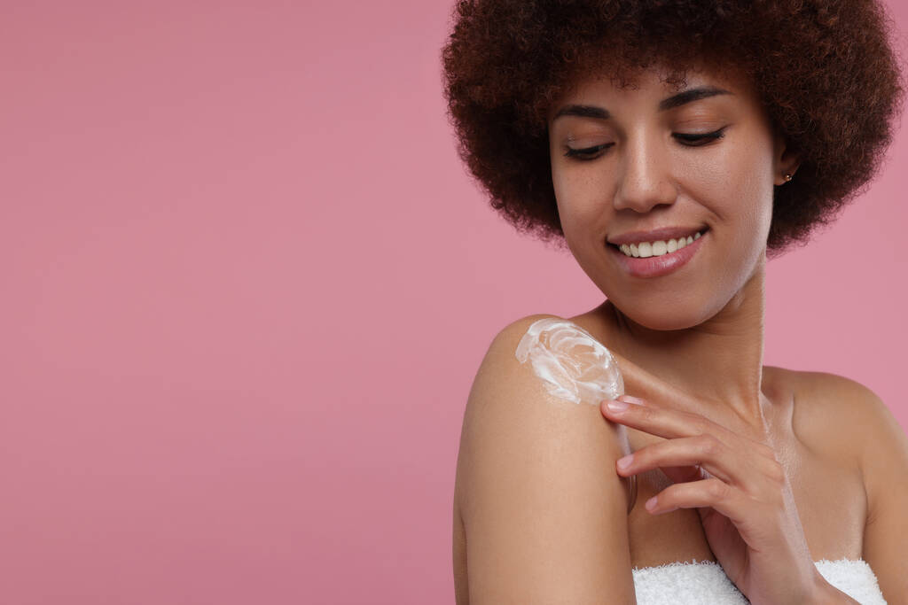Hermosa mujer joven aplicando crema corporal en el hombro sobre fondo rosa, espacio para el texto - Foto, Imagen