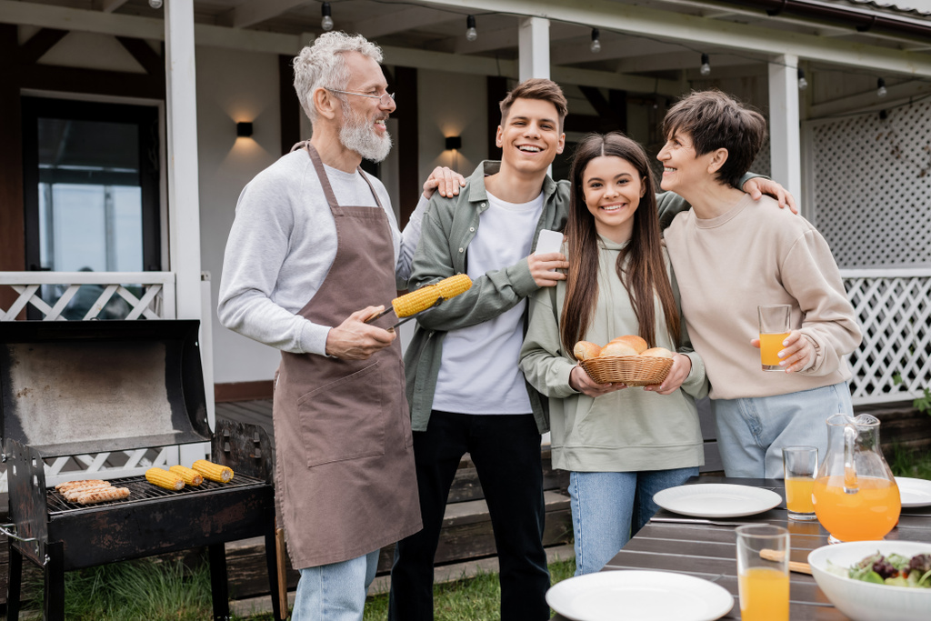 familiefoto, ouderdag, happy middle aged parents having bbq party with teenage daughter and young adult son, vader holding tangs with grilled corn, summer house, suburban life  - Foto, afbeelding