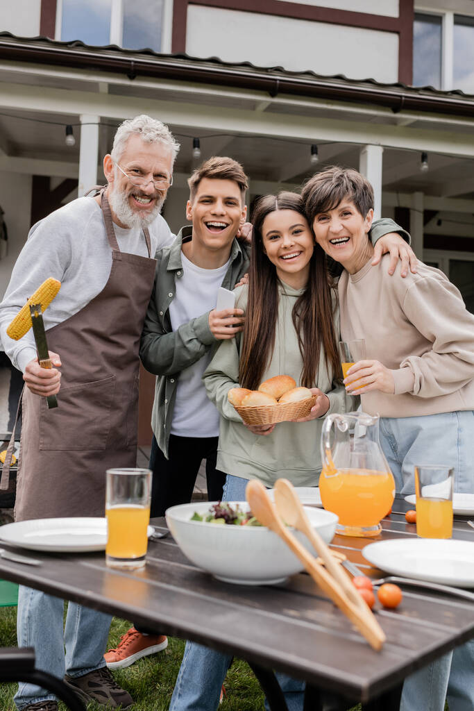 parents day, family photo, happy middle aged parents having bbq party with young adult son and teenage daughter, father holding tongs with grilled corn, summer house, suburban life  - Photo, Image