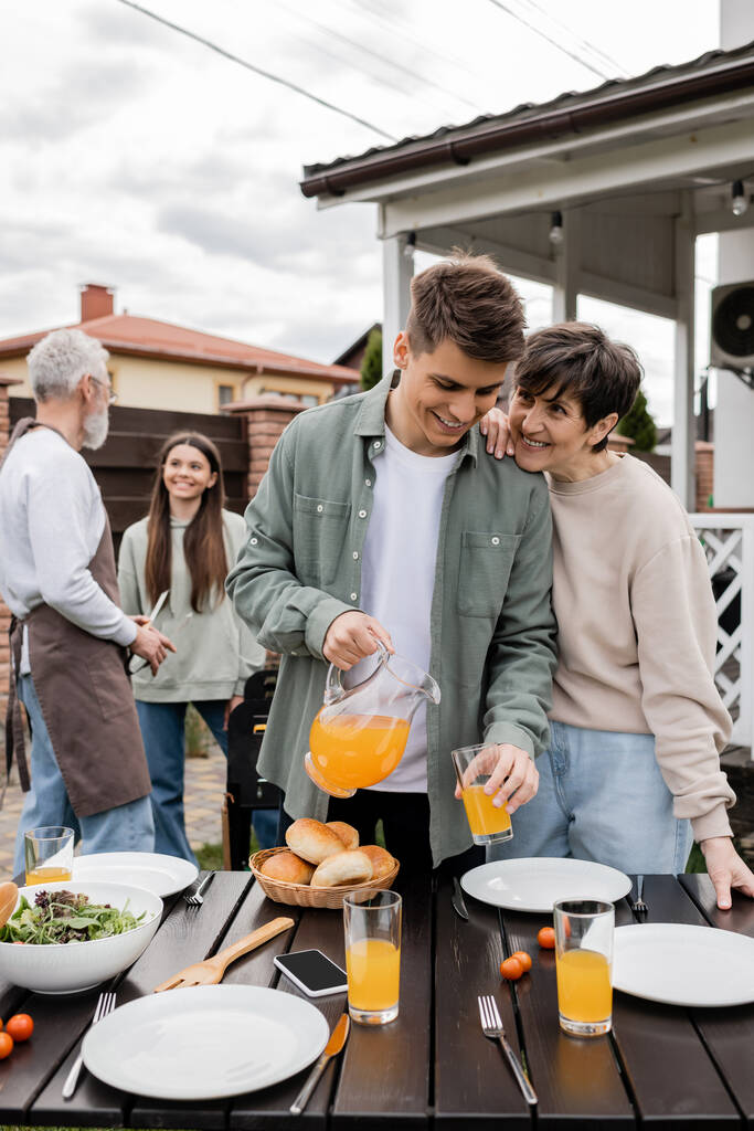 celebration of parents day, modern parenting, happy middle aged mother leaning on young adult son pouring orange juice in glass, father and daughter preparing food on bbq grill, summer, backyard  - Photo, Image