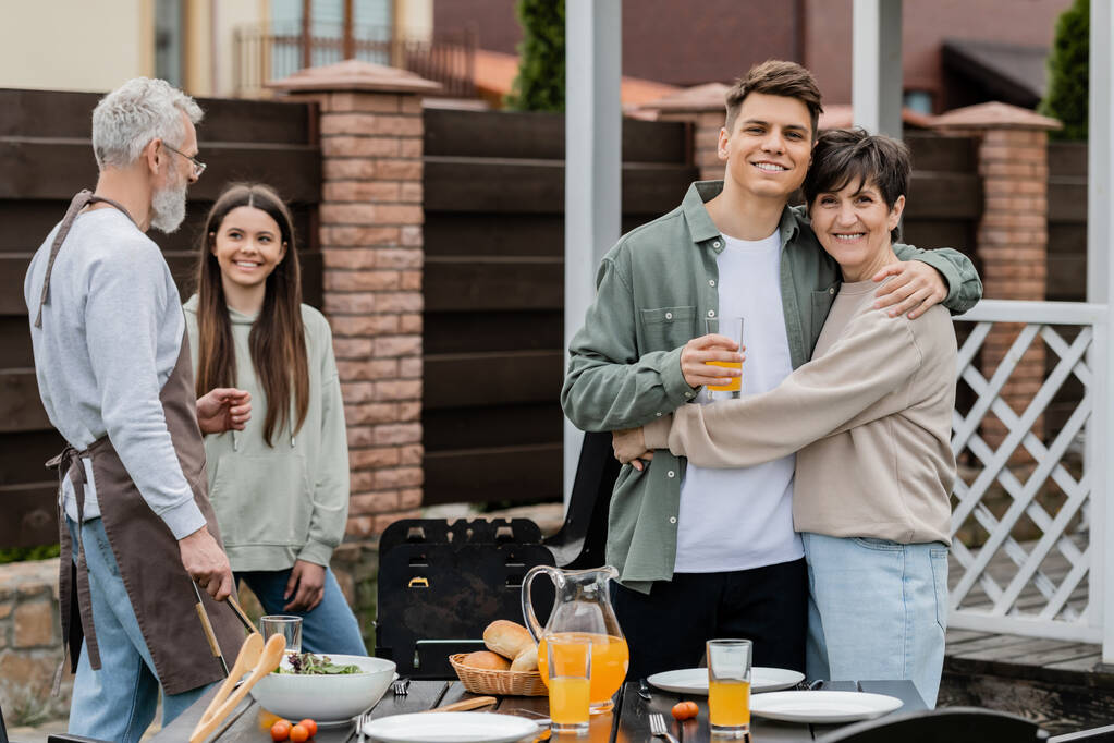 happy parents day, modern parenting, cheerful middle aged mother hugging young adult son holding glass of orange juice, father and daughter preparing food on bbq grill, summer, backyard, celebration  - Photo, Image