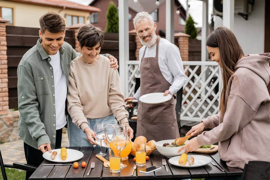 family bbq party, celebration of parents day, happy young adult son hugging middle aged mother, bearded father in apron looking at teenage daughter, backyard of family house  - Photo, Image