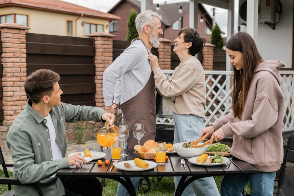 happy middle aged couple looking at each other during family bbq party, young adult son looking at cheerful parents, love, joyful teenage girl mixing salad, happy parents day, backyard, summer  - Photo, Image