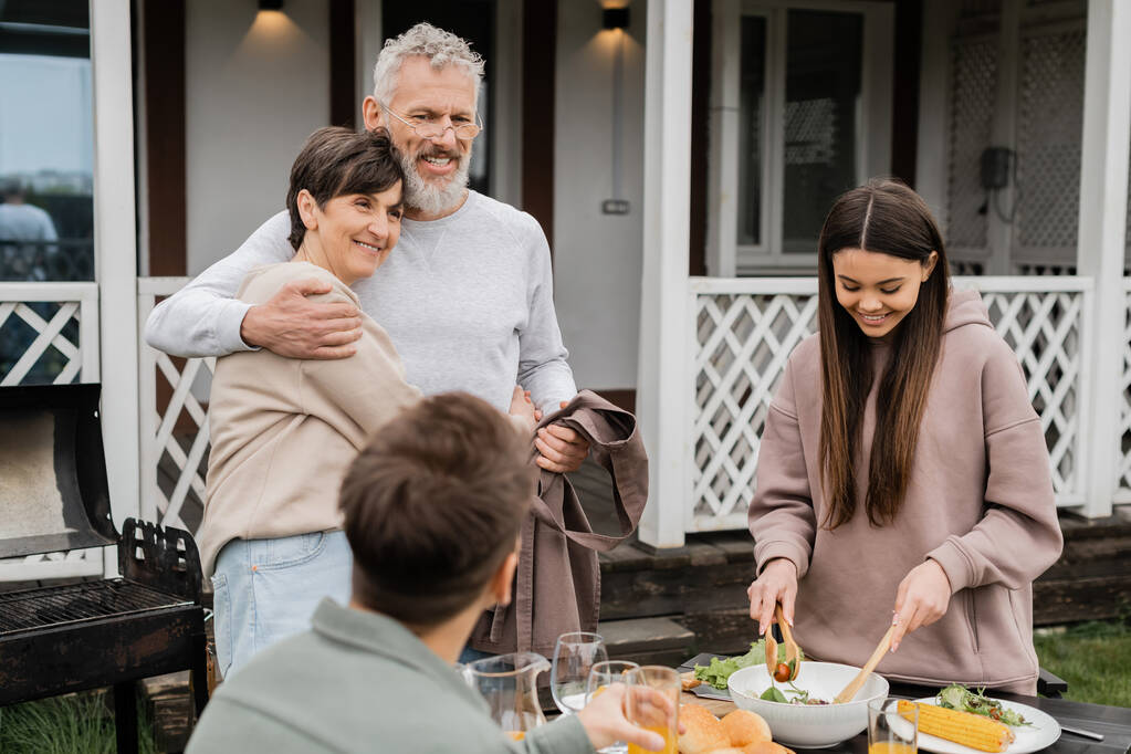 celebrating parents day, cheerful middle aged couple hugging near joyful teenage daughter serving salad next to adult brother, love, family grill party, summer, happy marriage, summer house backyard  - Photo, Image