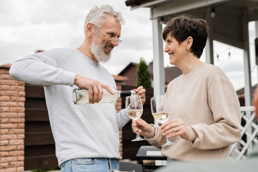 happy marriage, cheerful and bearded middle aged man in glasses pouring white wine into glass of joyful wife, backyard of summer house, romance, casual attire, spending time together  - Photo, Image