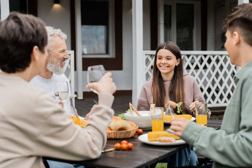 happy teenage girl looking at cheerful parents during family celebration, sitting on backyard of summer house, spending time together, eating grilled bbq food, happy parents day concept  - Photo, Image