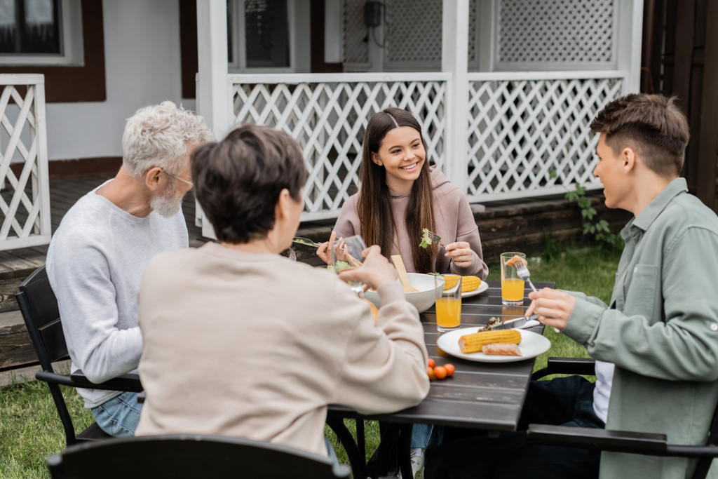 boldog tizenéves lány néz adutl testvér alatt családi ünnepség, ül a hátsó udvaron nyaralóház, időt töltenek együtt, eszik grillezett bbq élelmiszer, boldog szülők nap koncepció  - Fotó, kép