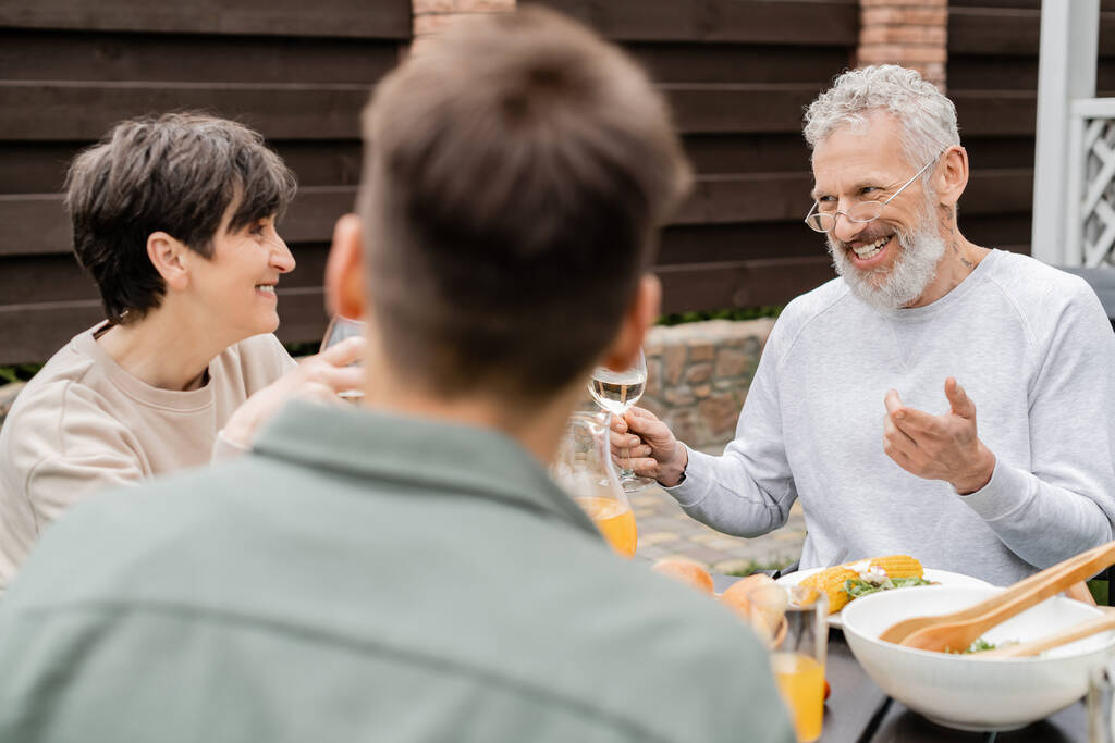 cheerful middle aged man looking wife near adult son and gesturing during bbq party, blurred foreground, backyard of summer house, spending time together, eating grilled bbq food, parents day concept  - Photo, Image