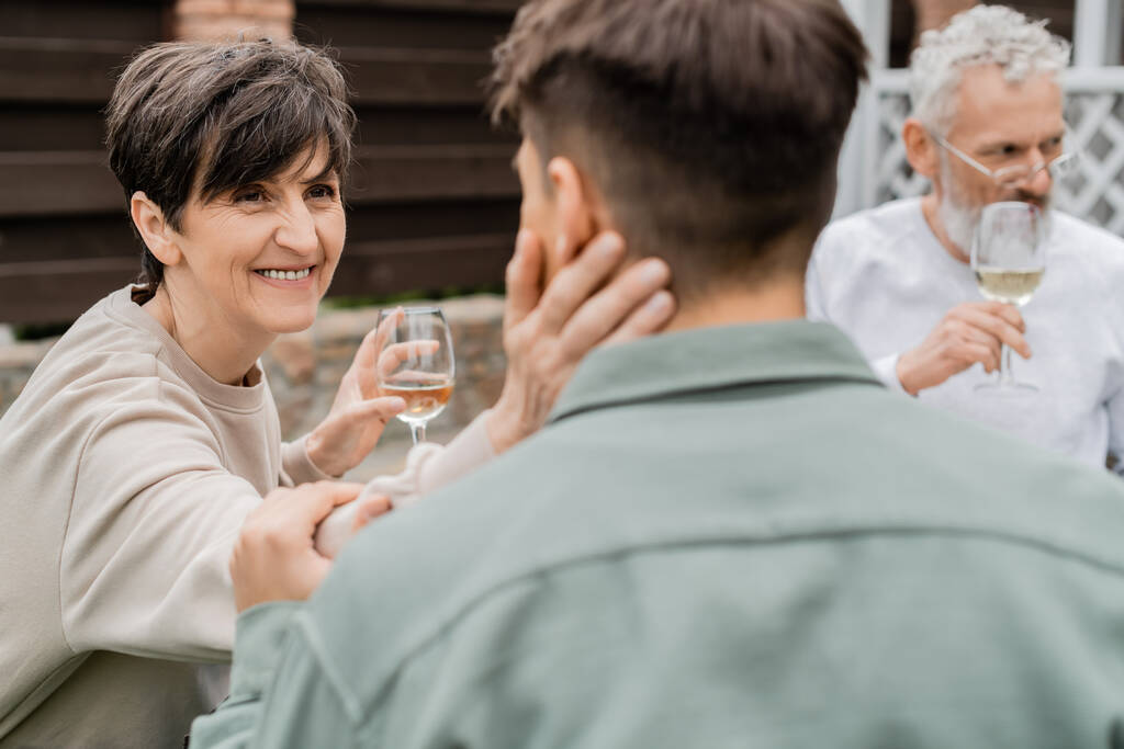 Smiling middle aged mother holding glass of wine and touching blurred adult son near husband at background during parents day celebration at backyard, family love and unity concept - Photo, Image