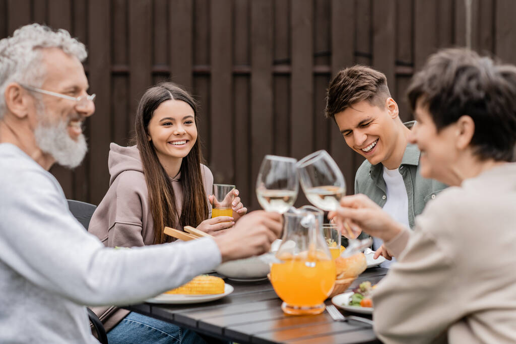 Smiling siblings sitting near tasty bbq food and blurred middle aged parents toasting with wine during parents day celebration at backyard, family love and unity concept, tradition and celebration - Photo, Image