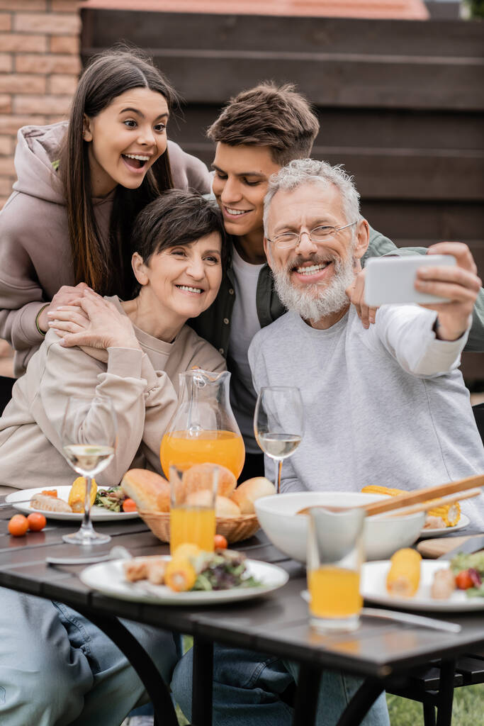 Позитивный зрелый отец делает селфи на смартфоне с женой и детьми возле размытой летней еды во время барбекю-вечеринки и празднования родительского дня на заднем дворе, счастливая концепция Дня родителей - Фото, изображение