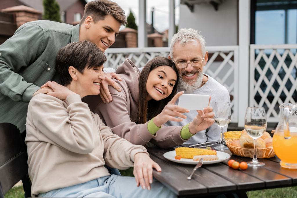 Positive teenage girl using smartphone near mature parents and father during bbq party with summer food and parents day celebration at backyard in june, happy parents day concept, special occasion - Photo, Image