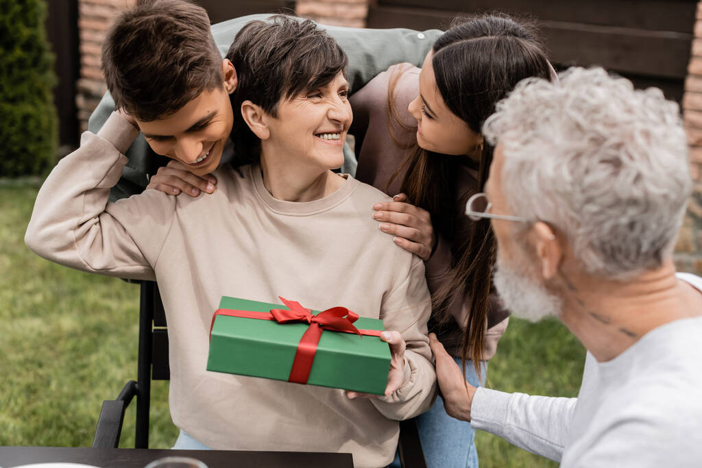 Pozytywne dzieci przytulające matkę w średnim wieku trzymającą pudełko prezentów w pobliżu rozmytego męża, spędzając czas podczas uroczystości Dnia Rodzica na podwórku w czerwcu, pielęgnując koncepcję więzi rodzinnych - Zdjęcie, obraz
