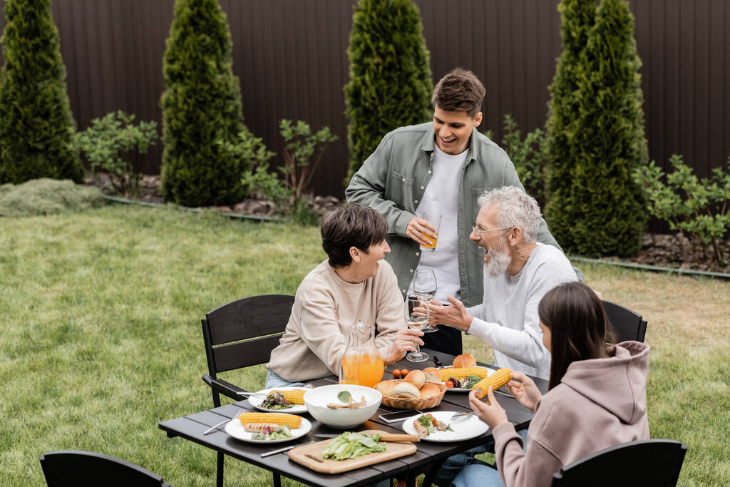 Aufgeregte und gut gelaunte Eltern mittleren Alters stoßen bei Grillparty im Hinterhof mit Weingläsern und sommerlichem Essen an, pflegen Familienbande, verbringen Zeit miteinander - Foto, Bild