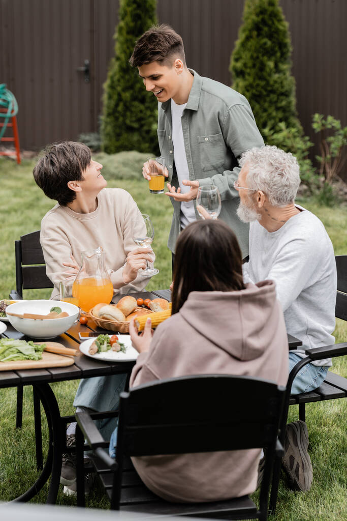 Веселый молодой человек, держащий апельсиновый сок и разговаривающий с матерью средних лет возле семьи и летняя еда барбекю во время празднования дня родителей на заднем дворе, лелея концепцию семейных уз - Фото, изображение