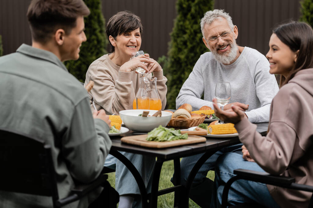 Pozytywni rodzice w średnim wieku patrząc na rozmazane dzieci w pobliżu letniego jedzenia podczas przyjęcia z grillem i dzień rodziców uroczystości na podwórku w czerwcu, kochający koncepcję obligacji rodzinnych - Zdjęcie, obraz