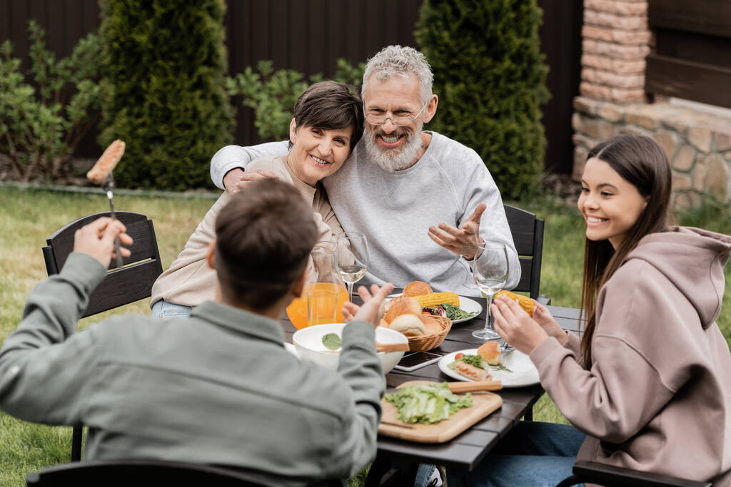 BBQパーティーや6月の裏庭での親の日のお祝い中に、中年の両親が夏の食べ物の近くの子供たちと抱き合って話し合って笑って、家族の絆の概念、特別な機会を大切にしています - 写真・画像