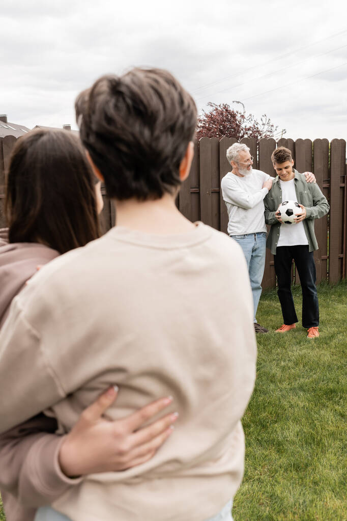 Батько середнього віку обіймає молодого сина з футболом біля розмитої сім'ї на задньому дворі під час святкування Дня батьків у червні, якісний час з концепцією батьків, особливий випадок
 - Фото, зображення