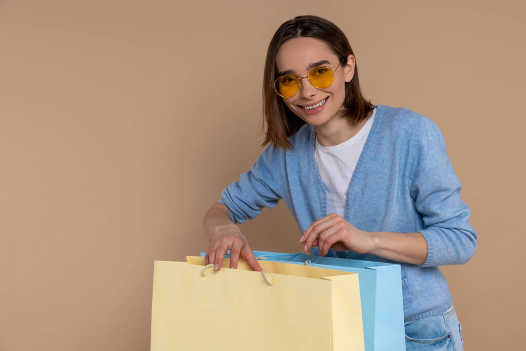 Zufriedene lächelnde Frau in lässiger Kleidung mit Einkaufstaschen vor beigem Hintergrund. - Foto, Bild