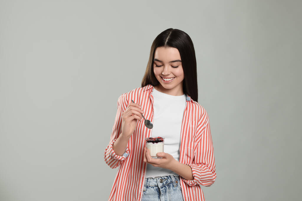 Ευτυχισμένο έφηβο κορίτσι με νόστιμο γιαούρτι και κουτάλι σε ανοιχτό γκρι φόντο. Χώρος για κείμενο - Φωτογραφία, εικόνα