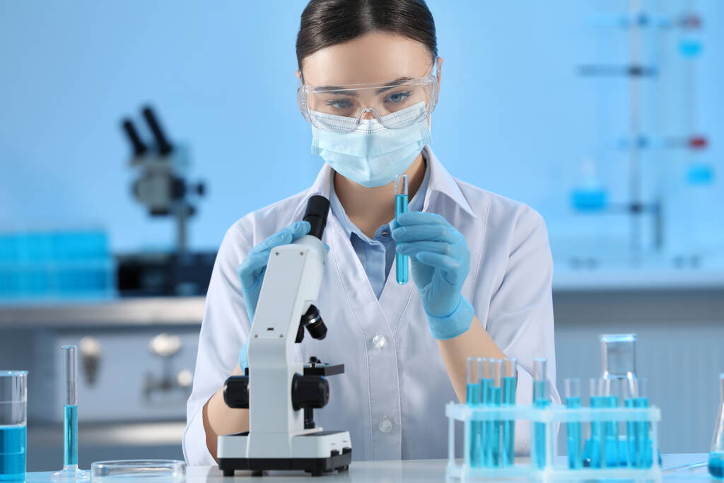 Scientifique tenant une éprouvette avec un liquide bleu clair et un microscope en laboratoire - Photo, image