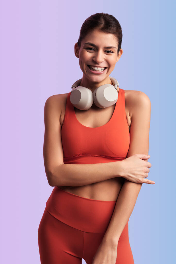 Pozitivní mladá žena sportovec v oranžové horní a podprsenka s bezdrátovými sluchátky s úsměvem a při pohledu do kamery - Fotografie, Obrázek