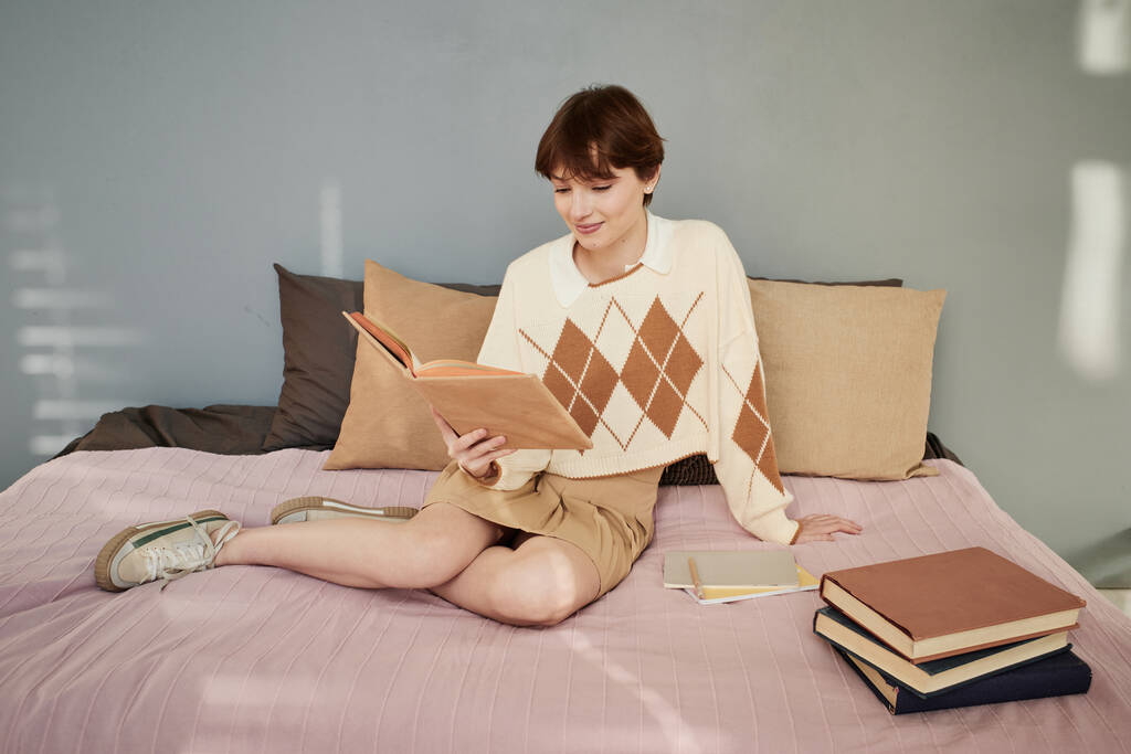 Περιεχόμενο θηλυκό φοιτητής πανεπιστημίου σε πουλόβερ κάθεται με το βιβλίο στο κρεβάτι στο δωμάτιο των μαθητών - Φωτογραφία, εικόνα