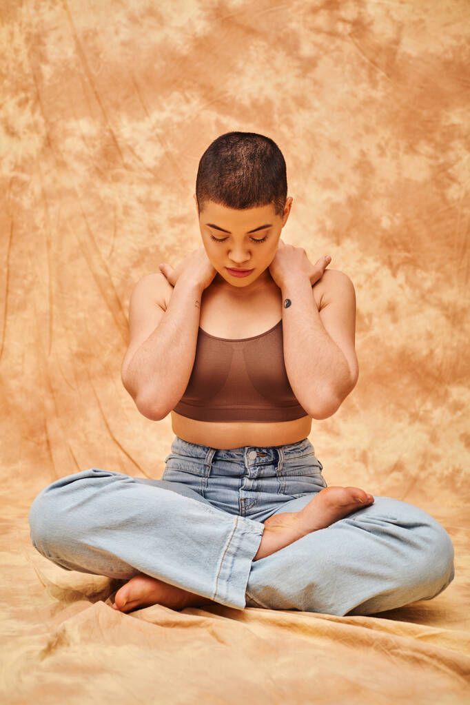 body confidence, relaxation, curvy young and tattooed woman in jeans and crop top sitting with crossed legs on mottled beige background, personal style, self-acceptance, generation z, denim fashion  - Photo, Image