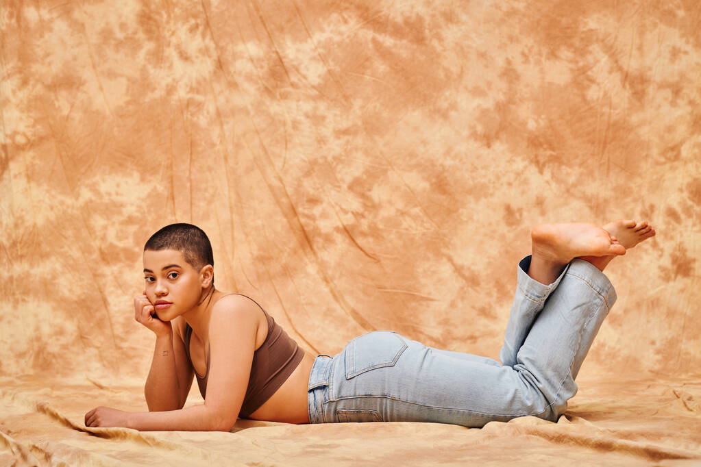 положительность тела, представление тела, кривая молодая и татуированная женщина в джинсах и топе лежа на пестром бежевом фоне, глядя на камеру, джинсовой моды, личный стиль, поколение z  - Фото, изображение