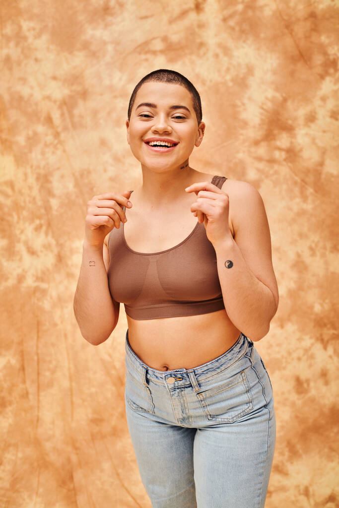 body positivity, happy and tattooed woman in crop top and jeans posing on mottled beige background, looking at camera, representation of body, different body shapes, generation z, youth, tattooed - Photo, Image