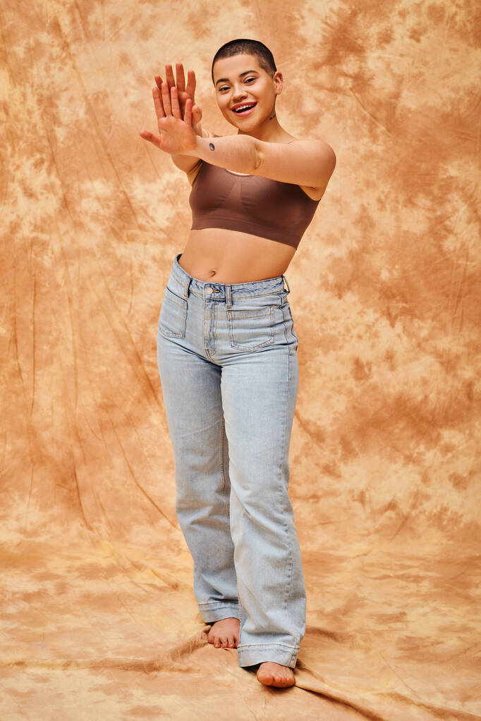 Body Positivity Bewegung, Jeans-Look, kurvige und fröhliche Frau im bauchfreien Top posiert mit ausgestreckten Händen auf fleckigem beigem Hintergrund, lässige Kleidung, Selbstakzeptanz, Generation z, tätowiert  - Foto, Bild