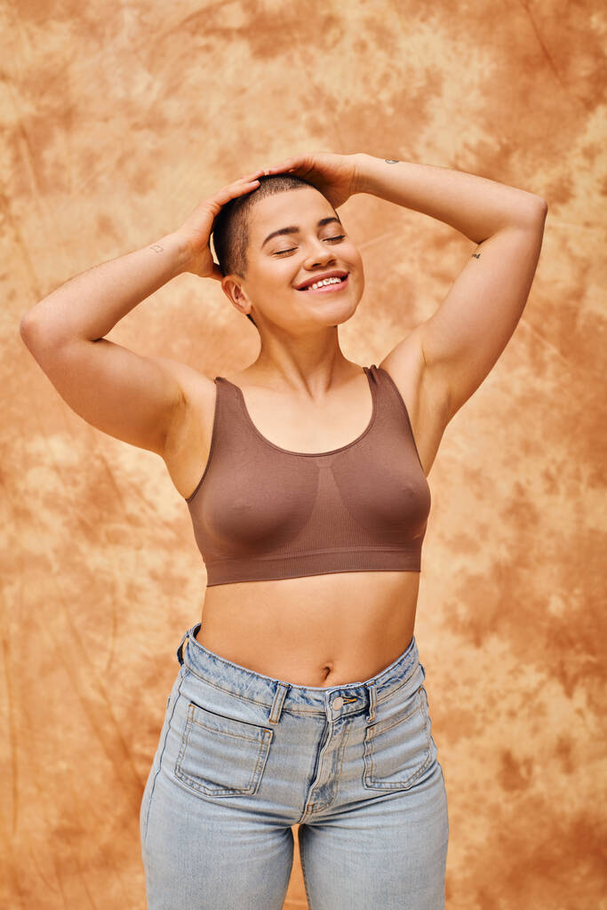 body acceptance, curvy and cheerful woman in crop top posing on mottled beige background, smiling with closed eyes, representation of body, different shapes, generation z, youth, tattooed   - Photo, Image