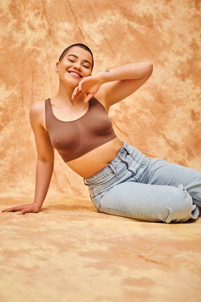 body love, jeans look, curvy and tattooed woman in casual attire sitting on mottled beige background, confidence, self-acceptance, generation z, body diversity, pretty and positive  - Foto, imagen